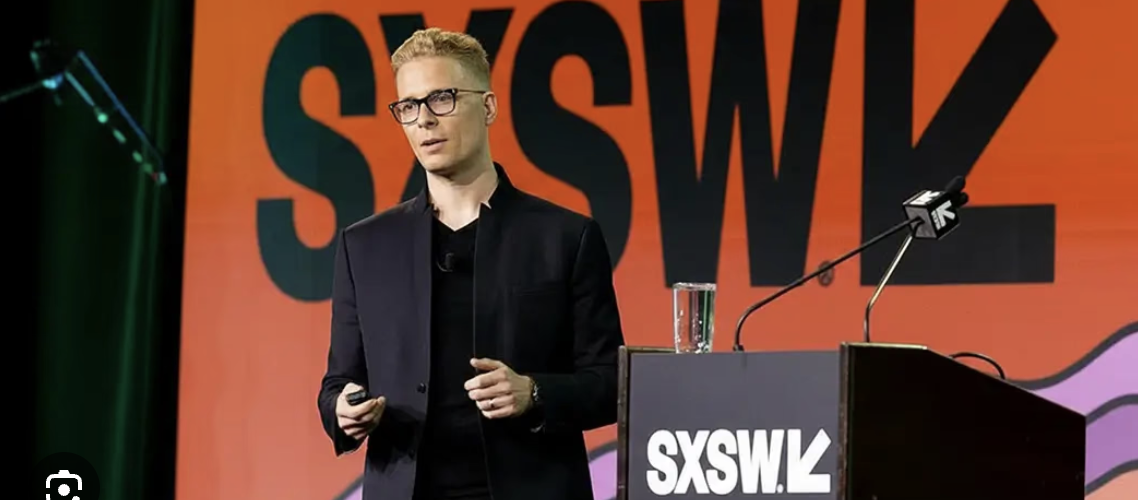 "Fotografia de Ian Beacraft palestrando no palco principal do SXSW. Ele veste um traje preto moderno e óculos, posicionado atrás de um púlpito com a logo do SXSW. Ao fundo, um grande telão laranja vibrante exibe a marca oficial do evento South by Southwest