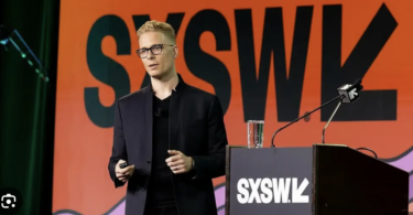 "Fotografia de Ian Beacraft palestrando no palco principal do SXSW. Ele veste um traje preto moderno e óculos, posicionado atrás de um púlpito com a logo do SXSW. Ao fundo, um grande telão laranja vibrante exibe a marca oficial do evento South by Southwest