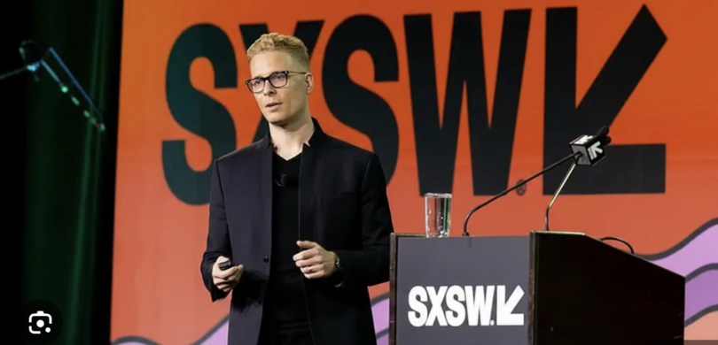 "Fotografia de Ian Beacraft palestrando no palco principal do SXSW. Ele veste um traje preto moderno e óculos, posicionado atrás de um púlpito com a logo do SXSW. Ao fundo, um grande telão laranja vibrante exibe a marca oficial do evento South by Southwest